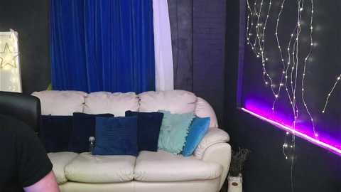 Video of a modern living room with a beige leather couch adorned with blue and turquoise pillows. The background features dark walls and a purple LED strip light, along with blue curtains.