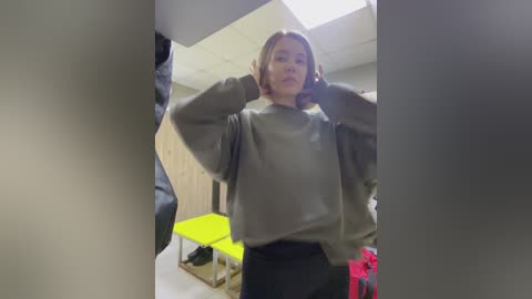 Media: Video of a young woman with light skin and shoulder-length blonde hair, wearing a grey sweater and black pants, posing in a brightly lit room with neon green chairs and wooden walls.