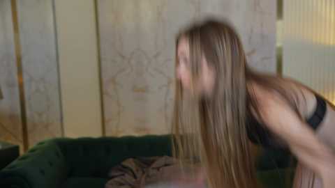 Media: A blurry video of a young woman with long, straight blonde hair, wearing a black bra, bending over in a dimly lit room with a green couch and beige curtains.