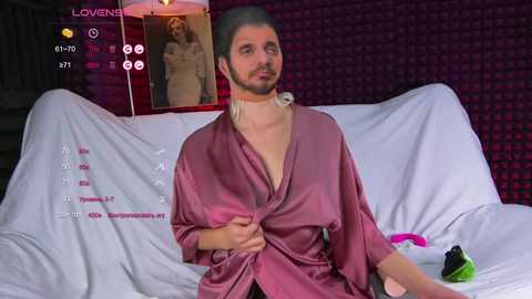 Video of a man with light skin and long, grey hair tied back, wearing a pink satin robe, sitting on a white bed in a dimly lit room.