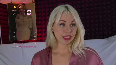 Video of a blonde woman in a pink satin robe, seated against a dark brick wall, with a live stream video of herself displayed in the background.