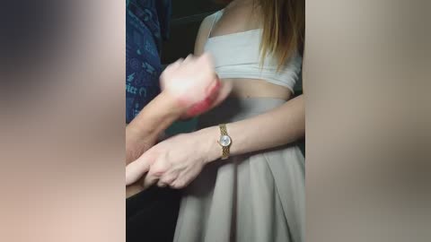 A video depicts a young woman in a white crop top and skirt, holding a pink, erect penis. She wears a gold wristwatch. The background is blurred.