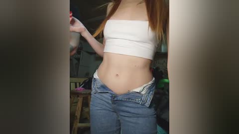Video of a slim, fair-skinned woman with long, straight, reddish-brown hair, wearing a white tube top and blue jeans, partially unbuttoned, in a dimly lit room with wooden furniture and cluttered background.