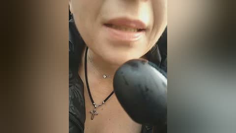 Close-up video of a woman's smiling face, partially obscured by a black microphone, wearing a black leather jacket and a necklace with a crucifix pendant.