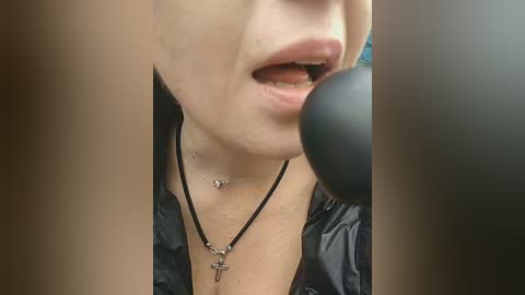 Video of a close-up of a woman's face, partially obscured by a microphone, wearing a black jacket, a silver cross necklace, and a black choker.