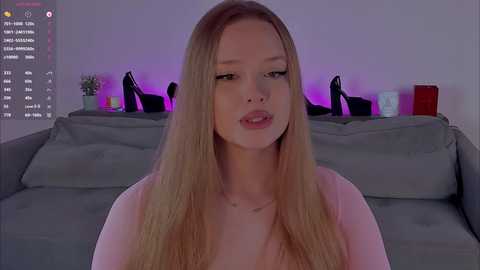 Video of a young, fair-skinned, blonde woman with long hair, wearing a pink top, sitting on a gray couch in a dimly lit room with purple lighting and black high heels on the couch behind her.