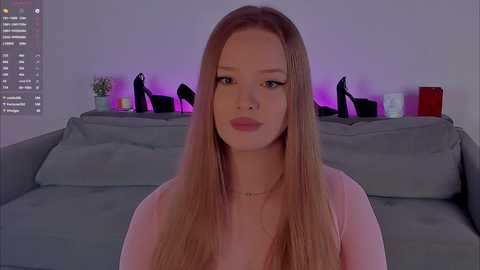 Video of a young, light-skinned woman with long, straight, blonde hair, wearing a pink top, sitting on a gray couch in a dimly lit room.