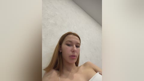 Media: Video of a young, fair-skinned woman with straight, shoulder-length red hair, wearing a white off-shoulder top, standing against a textured white wall. Her expression is neutral.