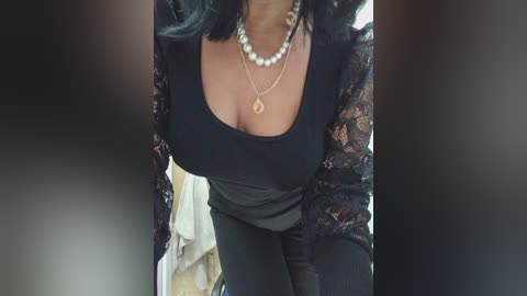 A video of a woman with medium-dark skin, wearing a low-cut black lace top revealing ample cleavage, pearl necklace, and a sheer black lace shawl. Background is blurred.