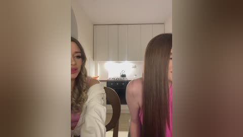 Media: Video of two young women in a modern kitchen. One has long wavy blonde hair, wearing a white jacket, and the other has long straight brown hair, wearing a pink top.