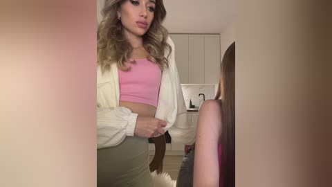 Media: A video of a curvy, fair-skinned woman with wavy blonde hair, wearing a pink crop top and khaki skirt, standing in a modern, minimalistic kitchen.