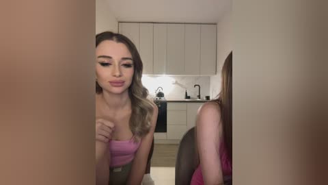 Media: A video of a young woman with light skin, long wavy blonde hair, and wearing a pink strapless top, standing in a modern, sleek kitchen with white cabinets and marble countertops.