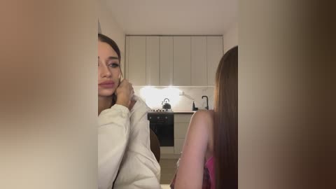 Media: Video of a young woman with fair skin and long, straight brown hair, wearing a white sweater, posing seductively in a modern kitchen with white cabinets and a stainless steel sink.