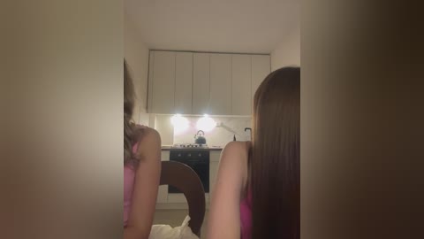 Media: Video of a woman with long brown hair, wearing a pink tank top, sitting in a dimly lit kitchen, seen from behind, with a mirror reflection.