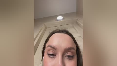 A close-up video of a young woman's face with long, straight brown hair, focused on her eyes and eyebrows, taken from a low angle. She has fair skin and wears subtle makeup. The background shows a beige wall with a white light fixture.