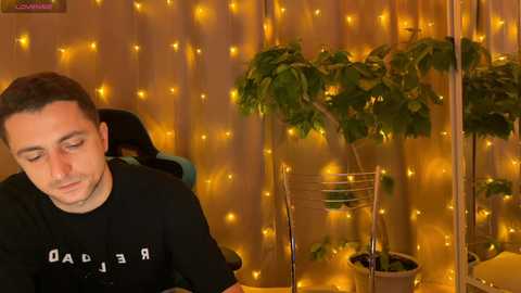 Media: A video of a man with short dark hair, wearing a black t-shirt, sitting in a chair with a plant beside him. The background features golden fairy lights and a potted plant, creating a cozy ambiance.