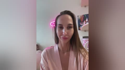 Video of a young woman with light skin and long brown hair, wearing a pink satin robe, smiling. Neon heart light illuminates background with colorful art and a bed.