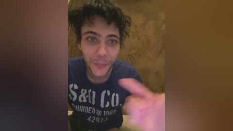 Video of a young man with curly hair, wearing a dark blue \"SBC\" t-shirt, smiling, pointing at the camera, against a beige, patterned wall background.