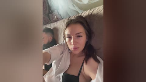 Media: Video of a woman with fair skin, brown hair, and light makeup, wearing a white shirt and black bra, sitting on a beige couch, taking a selfie.