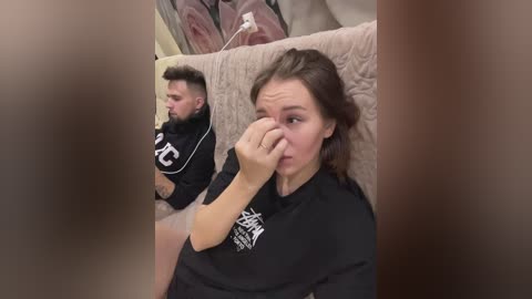 Media: Video of a man with a buzz cut and beard, wearing a black hoodie, sitting next to a woman with shoulder-length brown hair, also in a black hoodie. They are in a beige-carpeted room with a large mirror reflecting them.