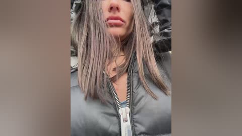 Media: Video of a woman with shoulder-length, straight, silver hair, wearing a black, quilted puffer jacket, partially zipped, with a neutral background.