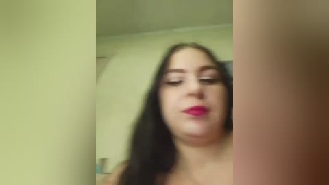 A blurred video of a young woman with fair skin, long dark hair, and bright red lipstick, wearing a sleeveless top, standing in a dimly lit room with yellow walls.