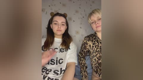 Media: Video of two young women in casual attire, one with a white t-shirt, the other in leopard print, standing against a patterned wall, with blurred faces in the foreground.