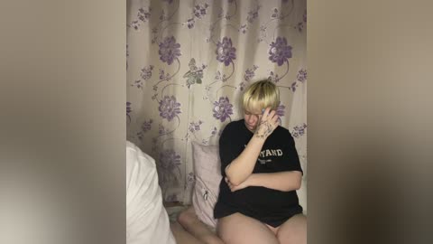 Media: Video of a blonde, plus-sized woman wearing a black t-shirt and no pants, sitting on a bed, covering her face with her hands, in a dimly lit room with floral-patterned curtains.