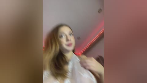 Media: A blurry video of a young woman with long, light brown hair, wearing a white shirt, standing in a dimly lit room with red LED strip lights on the ceiling.