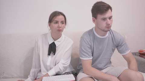 Video of a young woman with light brown hair in a white blouse and black tie, and a young man with short brown hair in a grey T-shirt and shorts, both sitting on a beige couch, looking bored.