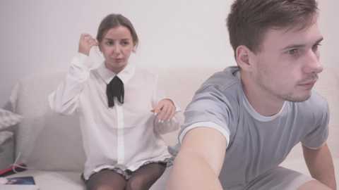 Video of a white woman in a white blouse and black bow tie, frustrated, gesturing at a white man in a gray t-shirt, sitting on a beige couch.