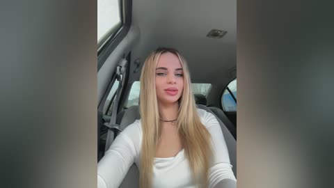 Media: Video of a young woman with long, straight blonde hair and fair skin, wearing a white long-sleeve top, sitting in a car.