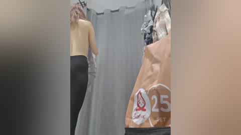 Media: Video of a woman with long, wavy hair, wearing black leggings and a yellow top, standing in a dressing room with beige curtains and a bag labeled \"25% off.\