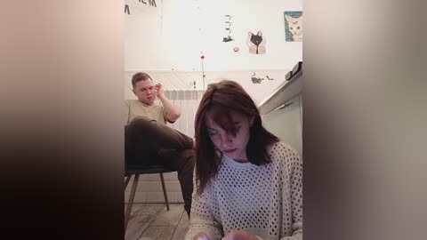 Media: Video of a young woman in a white knit sweater sitting on a tiled floor, looking down, while a young man sits on a chair in a dimly lit bathroom with white tiles and various decorations.