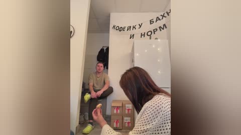 Media: Video of a man sitting in a room with beige walls, wearing a beige shirt and green shoes, while a woman with brown hair holds a phone. Boxes and a whiteboard with Russian text are visible.