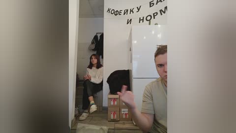 Media: Video of a woman in a white sweater sitting on a chair, looking at a mirror, with a man in the foreground wearing a beige shirt. The room has a white wall with Cyrillic writing, and boxes on the floor.