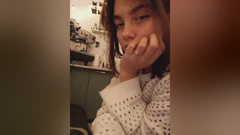 Media: A video of a young woman with medium skin tone and shoulder-length brown hair, wearing a white, knitted sweater, biting her finger. Background features a cluttered shelf with books, a green cabinet, and a wall with photos.