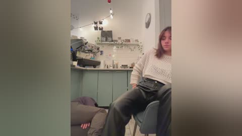 Media: Video of a young woman with shoulder-length brown hair, wearing a cream crochet sweater and dark jeans, sitting on a gray chair in a dimly lit kitchen with white cabinets and string lights.