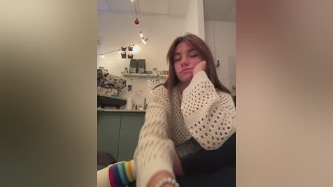 Media: A video captures a woman with long brown hair, wearing a white knitted sweater, sitting with her head resting on her hand, in a cozy, dimly lit kitchen.