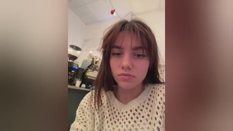 Media: Video of a young woman with long, dark hair and a neutral expression, wearing a white, knitted sweater. She sits in a dimly lit, cluttered kitchen with appliances and utensils visible in the background.