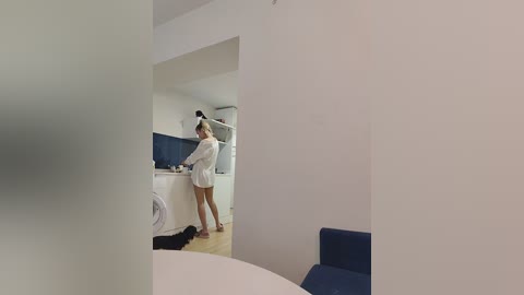 Media: Video of a modern, minimalist living room with a woman in a white robe, standing at a kitchen sink. The room features white walls, a blue-tiled backsplash, and a dark blue sofa.