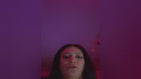 A video with a purple-pink tint featuring a young woman with long dark hair, fair skin, and light makeup, facing the camera with a neutral expression. The background is a plain, dark-colored wall.
