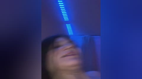A video of a woman with long, dark hair and a light skin tone, seen from the waist up, partially blurred. The background is dimly lit with blue LED lights, creating a moody, intimate atmosphere.