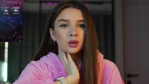 Media: A video of a young woman with long, straight brown hair, fair skin, and full lips, wearing a pink hoodie, touching her neck. Background shows a dimly lit room with blurred furniture.