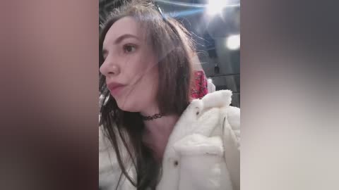 Video of a pale-skinned woman with long dark hair, wearing a white fur-trimmed jacket and a black choker, making a pouty face. Background features dimly lit, industrial-style metal pipes.