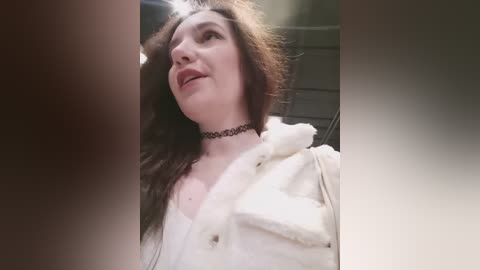 Video of a pale-skinned woman with long brown hair, wearing a black choker and white fur-trimmed jacket, looking upward with a serene expression, indoors with soft lighting.
