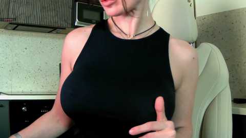 Media: A video of a fair-skinned woman with a slender build and medium-sized breasts, wearing a black sleeveless top, sitting in a modern kitchen with white countertops and a microwave. She has a black choker and her head is turned slightly, revealing her profile.