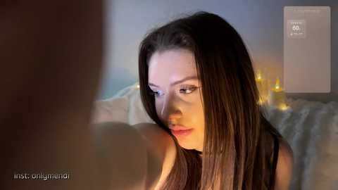 Video of a young, fair-skinned woman with long, dark hair, wearing a black top, looking introspective. Background features candles and a blurred bed.