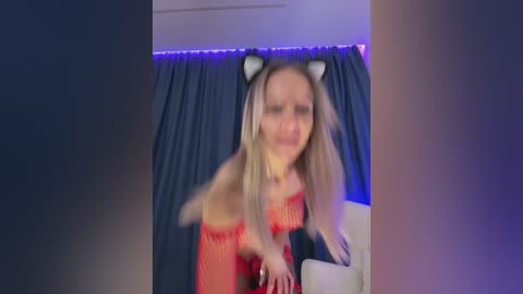 Media: Video of a blonde woman with cat ears, wearing a red and orange lingerie set, standing in a dimly-lit room with blue curtains and a purple light strip above.