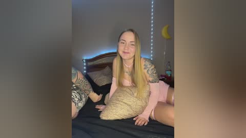 Media: Video of a young, light-skinned woman with long blonde hair, wearing a pink off-shoulder top and matching shorts, sitting on a bed with a black blanket. She has extensive tattoos on her arms. Background includes a wooden headboard and fairy lights.